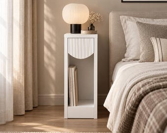 May include: A tall, narrow white bedside table featuring a single drawer with a vertical ribbed front panel and an open storage compartment below. The table holds a round, white, ribbed lamp and a small vase with dried flowers.