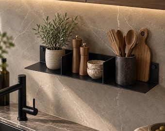 May include: A matte black metal wall shelf featuring three sections holding a potted green plant, two wooden pepper mills, two small speckled bowls, a dark gray utensil crock with wooden spoons, and a wooden cutting board.