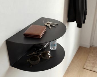 May include: A matte black semi-circular wall-mounted shelf with two tiers. The top tier holds a brown leather wallet and a set of metal keys. The bottom tier holds a pair of brown-tinted sunglasses, a black key fob, and a small clear plastic bottle.