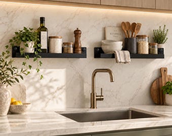 May include: Two black floating shelves mounted on a marble wall above a kitchen sink. The shelves hold a glass bottle, ceramic jars, a wooden pepper mill, a book titled The Kinfolk Table, wooden spoons, and small potted plants.