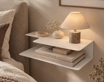 May include: A white metal floating wall shelf with two tiers. The top tier holds a small beige vase with dried flowers, a round dish, and a lamp with a marble base. The bottom tier holds two books titled The Kinfolk Home and At Home in the Modern World.