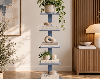 May include: A tall, blue, vertical shelving unit with six staggered shelves. The shelves hold a potted trailing plant, decorative ceramic objects, a candle, and several books with titles including The Kinfolk Home, A Life More Calm, and Loewe.
