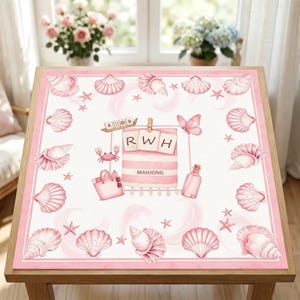 May include: A square pink and white table mat featuring a border of seashells and starfish. The centre displays a beach towel with the letters RWH, a tote bag, a bottle, a crab, a butterfly, and a mahjong tile set with the text MAHJONG.