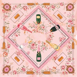 May include: A square scarf with a light pink background featuring a floral pattern of pink roses. The design includes five champagne bottles in various colors, the text "Nikita Mahjong Set", and border text reading "CRACKS-RED DRAGON BAM-GREEN DRAGON DOTS-SOAPS JOKERS MAY NOT BE USED IN SINGLES OR PAIRS".