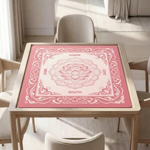 May include: A square mahjong mat featuring a pink and cream color scheme with a central lotus flower motif. The border displays intricate floral and cloud patterns, with the cardinal directions North, South, East, and West printed in capital letters.