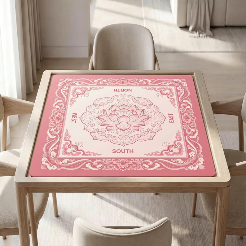 May include: A square mahjong mat featuring a pink and cream color scheme with a central lotus flower motif. The border displays intricate floral and cloud patterns, with the cardinal directions North, South, East, and West printed in capital letters.