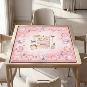 May include: A square pink mahjong mat featuring a border of various seashells and starfish. The center displays a beach-themed illustration with a striped towel labeled RWH, a blue crab, a tote bag, a seagull, and a message in a bottle.