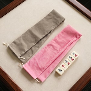 May include: A rectangular taupe fabric pouch and a rectangular pink fabric pouch, both featuring drawstring closures. Beside the pink pouch are four off-white mahjong tiles, each displaying black Chinese characters, with one tile featuring a red character.