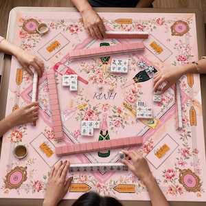 May include: A square mahjong mat featuring a light pink background with floral patterns, champagne bottle illustrations, and the text Nikita and RWH. The mat includes rows of pink mahjong tiles, white tiles with red and black characters, and dice.