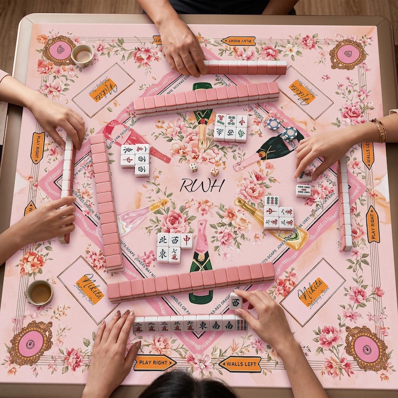 May include: A square mahjong mat featuring a light pink background with floral patterns, champagne bottle illustrations, and the text Nikita and RWH. The mat includes rows of pink mahjong tiles, white tiles with red and black characters, and dice.