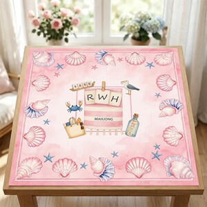 May include: A square pink mat featuring a border of various seashells and starfish. The center displays a striped towel with the letters RWH, a beach bag, a seagull, a message in a bottle, a blue crab, and a mahjong tile with the text MAHJONG.