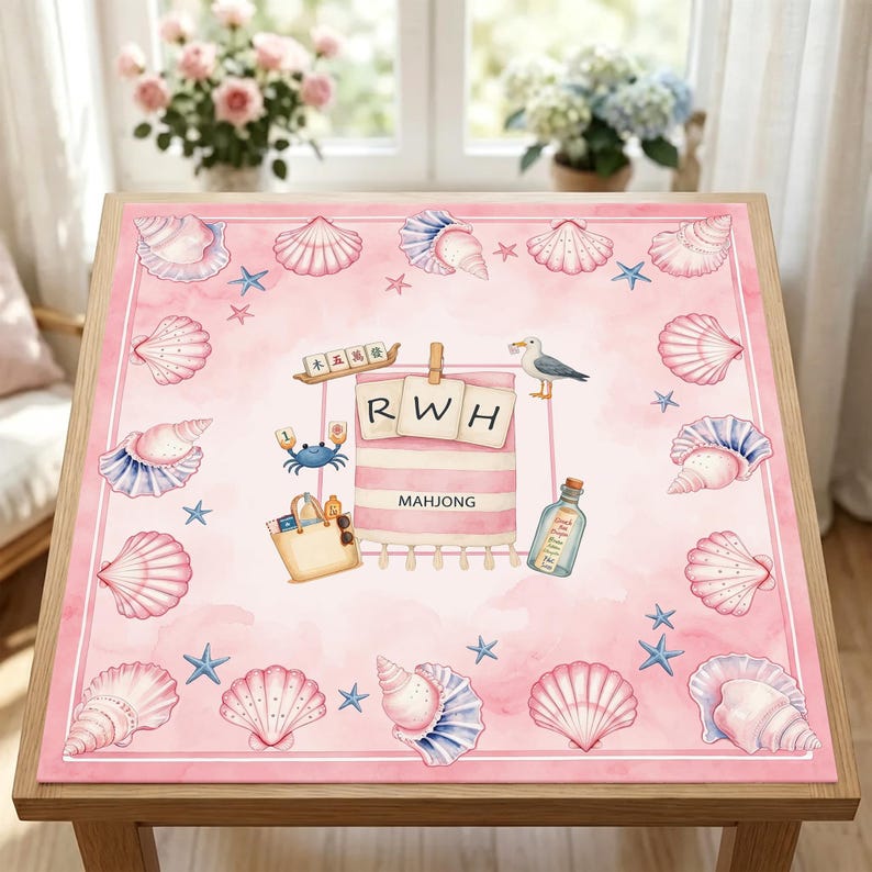 May include: A square pink mat featuring a border of various seashells and starfish. The center displays a striped towel with the letters RWH, a beach bag, a seagull, a message in a bottle, a blue crab, and a mahjong tile with the text MAHJONG.