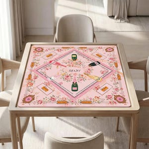 May include: A square pink game mat featuring a central diamond border, floral illustrations, and four champagne bottle graphics. The center displays the letters RWH, surrounded by text, game markers, and decorative frames in a soft pastel color palette.