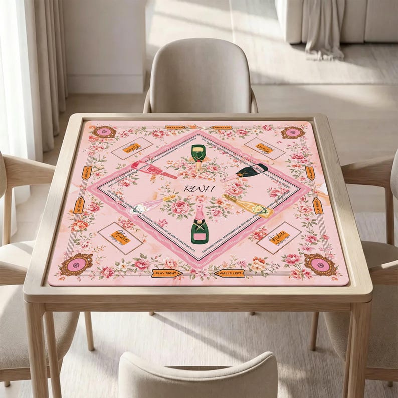 May include: A square pink game mat featuring a central diamond border, floral illustrations, and four champagne bottle graphics. The center displays the letters RWH, surrounded by text, game markers, and decorative frames in a soft pastel color palette.