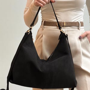 Peut inclure: Un sac &agrave; bandouli&egrave;re en daim noir avec une fermeture &eacute;clair et des accessoires dor&eacute;s. Le sac a une longue bandouli&egrave;re et un design minimaliste, adapt&eacute; &agrave; un usage quotidien. Le sac est tenu par une personne.
