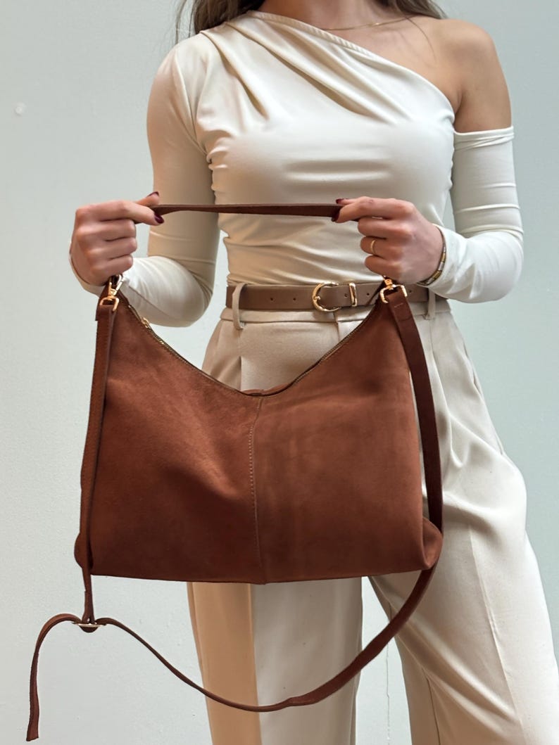 Peut inclure: Sac &agrave; bandouli&egrave;re en daim marron avec une longue bandouli&egrave;re r&eacute;glable et une poign&eacute;e plus courte. Le sac a une fermeture &eacute;clair et un design minimaliste. Le sac est tenu par une personne portant un haut blanc et un pantalon beige.