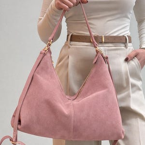 Peut inclure: Un sac &agrave; bandouli&egrave;re en daim rose poudr&eacute; avec une fermeture &eacute;clair et des accessoires dor&eacute;s. Le sac a une longue bandouli&egrave;re r&eacute;glable et un design souple de style sac hobo. Le sac est tenu par une personne portant un haut blanc et un pantalon beige.