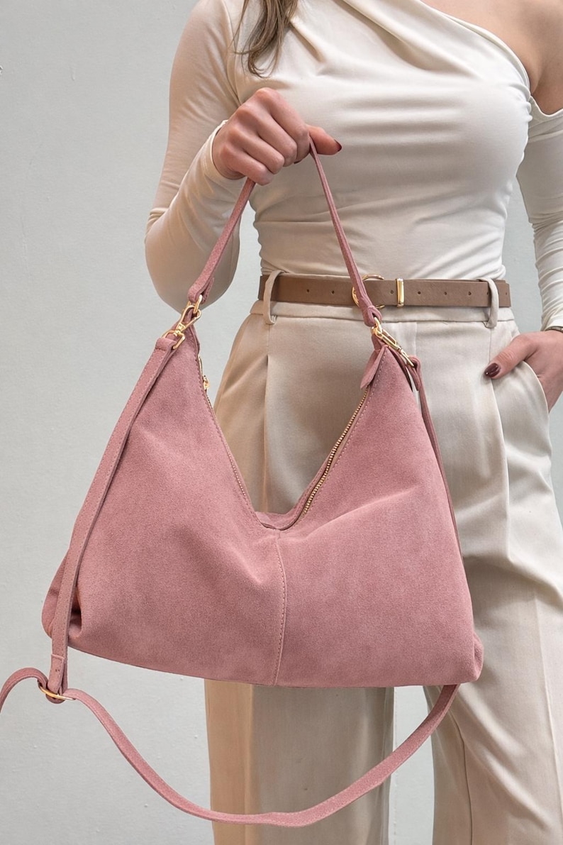Peut inclure: Un sac &agrave; bandouli&egrave;re en daim rose poudr&eacute; avec une fermeture &eacute;clair et des accessoires dor&eacute;s. Le sac a une longue bandouli&egrave;re r&eacute;glable et un design souple de style sac hobo. Le sac est tenu par une personne portant un haut blanc et un pantalon beige.
