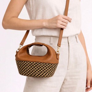 Pu&ograve; includere: Una borsa in pelle marrone con manico superiore arrotondato e tracolla staccabile. La parte inferiore della borsa presenta una trama intrecciata con un motivo nero e beige. La borsa ha finiture color oro che collegano la tracolla al corpo.