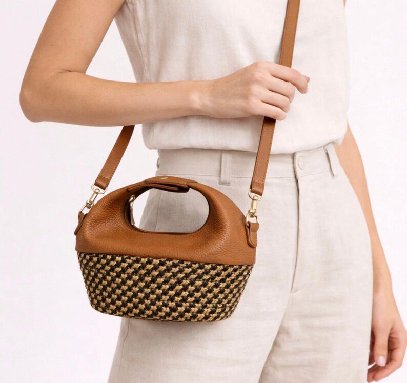 Pu&ograve; includere: Una borsa in pelle marrone con manico superiore arrotondato e tracolla staccabile. La parte inferiore della borsa presenta una trama intrecciata con un motivo nero e beige. La borsa ha finiture color oro che collegano la tracolla al corpo.