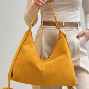Peut inclure: Sac &agrave; bandouli&egrave;re en daim jaune moutarde avec fermeture &eacute;clair. Le sac est dot&eacute; d'une longue bandouli&egrave;re r&eacute;glable et d'accessoires dor&eacute;s. La personne porte un haut blanc &agrave; manches longues et un pantalon beige.