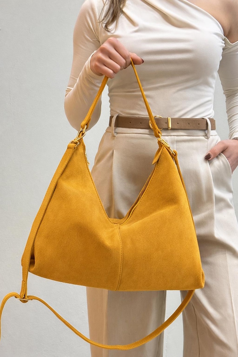 Peut inclure: Sac &agrave; bandouli&egrave;re en daim jaune moutarde avec fermeture &eacute;clair. Le sac est dot&eacute; d'une longue bandouli&egrave;re r&eacute;glable et d'accessoires dor&eacute;s. La personne porte un haut blanc &agrave; manches longues et un pantalon beige.
