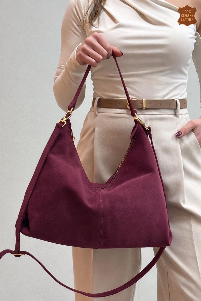 Peut inclure: Un sac &agrave; bandouli&egrave;re en daim bordeaux avec une fermeture &eacute;clair et des accessoires dor&eacute;s. Le sac a une longue bandouli&egrave;re et porte l'&eacute;tiquette "100% Cuir v&eacute;ritable".