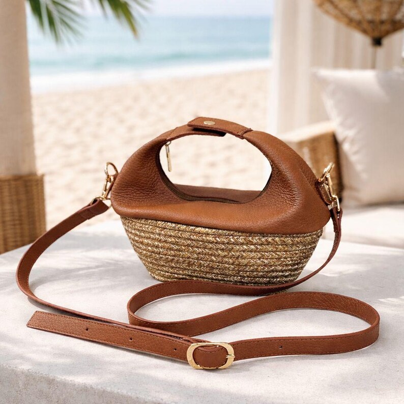 Pu&ograve; includere: Una borsa in pelle marrone e paglia intrecciata con tracolla. La borsa ha una forma arrotondata con un manico superiore e una fibbia color oro sulla tracolla. La borsa &egrave; appoggiata su una superficie bianca.