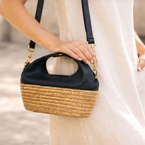 Pu&ograve; includere: Una borsa strutturata con una sezione superiore in pelle nera martellata e una base in paglia intrecciata. La borsa presenta un manico superiore, una tracolla rimovibile con dettagli in metallo dorato e una silhouette curva.
