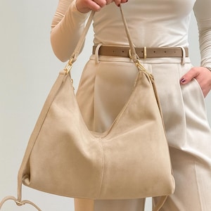Peut inclure: Un sac &agrave; bandouli&egrave;re en daim beige avec un design incurv&eacute;. Le sac a une longue bandouli&egrave;re r&eacute;glable et des accessoires dor&eacute;s. Le sac est tenu par une personne portant un haut et un pantalon cr&egrave;me.