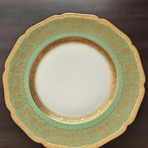 Può includere: Un piatto decorativo con bordo smerlato. Il piatto presenta un centro bianco, un bordo verde chiaro con un motivo floreale dorato e un bordo dorato. Il piatto è appoggiato su una superficie marrone scuro.