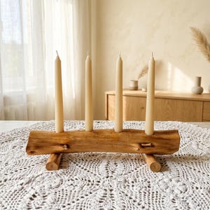 Könnte beinhalten: Ein Kerzenhalter aus einem natürlichen Holzstamm mit vier vertikalen zylindrischen Öffnungen, in denen jeweils eine lange, schlanke, hellbeige Stabkerze steckt. Der Stamm wird von zwei kleinen, horizontalen Holzdübeln als Standfüße gestützt.