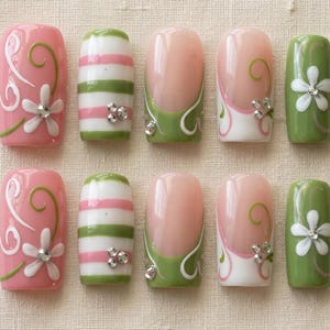 K&ouml;nnte beinhalten: Ein Set aus zehn k&uuml;nstlichen N&auml;geln in verschiedenen Designs. Die N&auml;gel zeigen rosa, gr&uuml;ne und wei&szlig;e Farben mit floralen und gestreiften Mustern. Einige N&auml;gel haben ein French-Tip-Design mit gr&uuml;nen Akzenten und Strasssteinen.