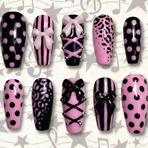 May include: A collection of ten artificial nails in various designs. The nails feature pink and black colors, with patterns including polka dots, stripes, and leopard print. Some nails are adorned with bows and decorative accents.