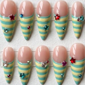 Op de afbeelding: Een set van tien amandelvormige press-on nagels met een nude basis en horizontale gele en blauwgroene strepen op de uiteinden. Elke nagel is versierd met kleine, kleurrijke stervormige en ronde strass-steentjes in rood, blauw en transparant.