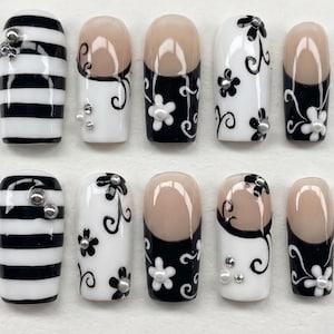 May include: A set of ten press-on nails featuring a black and white color palette. Designs include horizontal stripes, black floral patterns on white backgrounds, and black French tips with white flower accents. Small silver studs are applied to several nails.