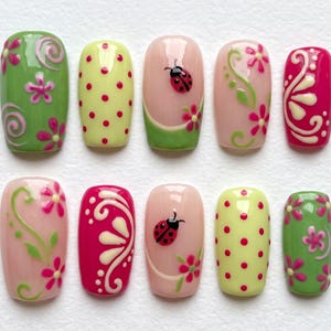 May include: A set of ten press-on nails featuring a variety of designs including light green with pink flowers and swirls, yellow with red polka dots, nude with ladybug and flower accents, and bright pink with white swirling floral patterns.