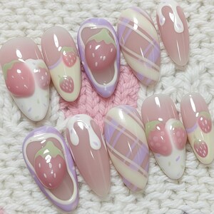May include: A set of ten artificial nails with a strawberry theme. The nails feature a variety of designs, including 3D strawberries, plaid patterns, and dripping white accents. The nails are in shades of pink, white, and purple.