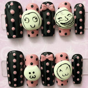 May include: A set of press-on nails with a playful design. The nails feature a combination of black and pink polka dots, with some nails adorned with cartoon faces and a pink bow. The nails are displayed on a clear plastic sheet.