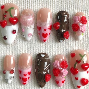 May include: A collection of ten artificial nails with various designs. Some nails feature cherries, hearts, and floral accents. The color palette includes white, pink, brown, and red. The nails are arranged on a white, textured surface with embroidered hearts and roses.