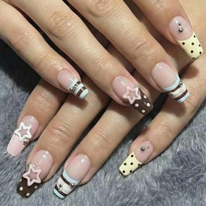 May include: A close-up of a set of long, decorated fingernails. The nails feature a variety of designs, including polka dots, stripes, and star accents. Colors include brown, pink, white, and light blue. Some nails have small silver studs.