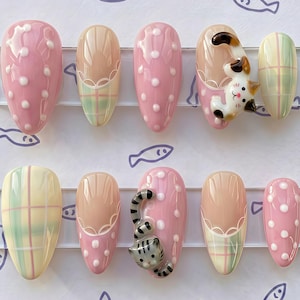 Pode incluir: Um conjunto de dez unhas artificiais com vários designs. As unhas apresentam uma base rosa claro com bolinhas brancas, padrões xadrez pastel e decorações de gatos 3D. As unhas são exibidas em um suporte de plástico transparente sobre um fundo branco com ilustrações de peixes.