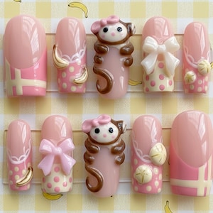 May include: A collection of ten artificial nails in pink, white, yellow, and brown. Some nails feature monkey designs, bows, and banana details. The background is a yellow and white checkered pattern with banana illustrations. The nails are designed for cosmetic use.