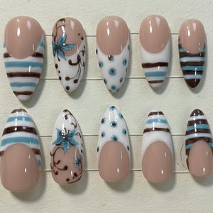 Pode incluir: Um conjunto de dez unhas artificiais com vários designs. Algumas unhas apresentam uma base nude com listras brancas, marrons e azuis. Outras têm uma base nude com bolinhas brancas e azuis ou padrões florais. As unhas são ovais.
