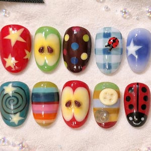 Pode incluir: Um conjunto de dez unhas artificiais com vários desenhos. As unhas apresentam desenhos como estrelas, fatias de maçã, bolinhas, xadrez e uma joaninha. As cores incluem vermelho, verde, azul, castanho e amarelo. As unhas têm formato oval.