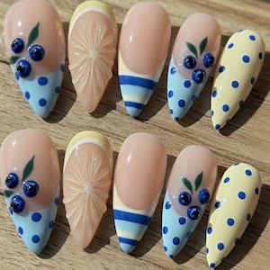 May include: A set of ten artificial nails with various designs. Some nails feature a light pink base with blue polka dots and blueberry accents. Others have a lemon slice design, blue stripes, or yellow polka dots. The nails are almond-shaped.