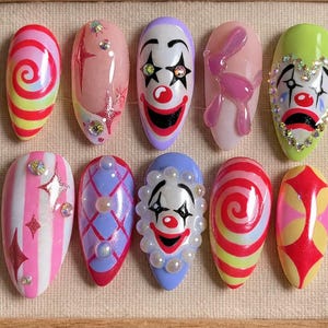 May include: A set of ten artificial nails with a clown theme. The nails feature various designs, including clown faces, spiral patterns, and decorative embellishments. Colors include red, pink, green, and purple.