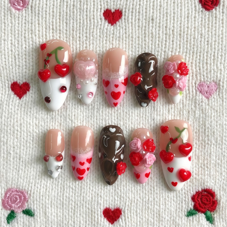 Op de afbeelding: Een set van tien kunstnagels met verschillende ontwerpen. Sommige nagels hebben kersen, harten en bloemaccenten. Kleuren zijn wit, roze, bruin en rood. De nagels zijn gerangschikt op een wit, getextureerd oppervlak met geborduurde harten.