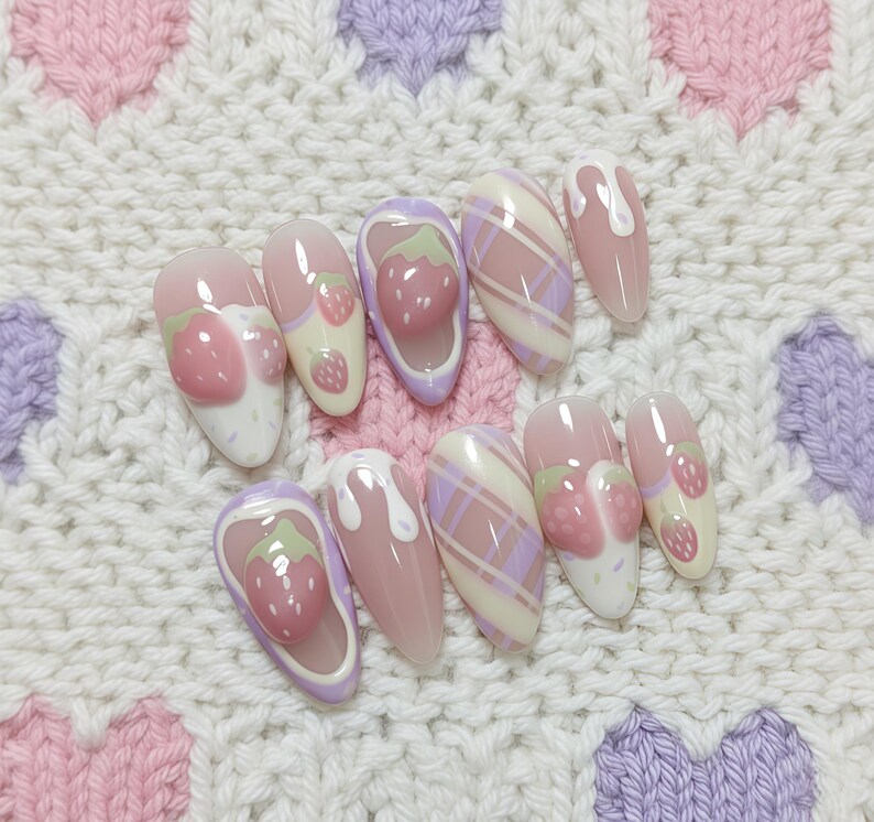 May include: A set of ten artificial nails with a strawberry theme. The nails feature a variety of designs, including 3D strawberries, plaid patterns, and dripping white accents. The nails are in shades of pink, white, and purple.