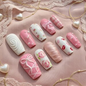 May include: A collection of artificial nails in various designs. The nails feature pink, white, and pearlescent colors with patterns like bows, roses, and lip prints. Some nails have pearl accents, and others have a textured swirl design.